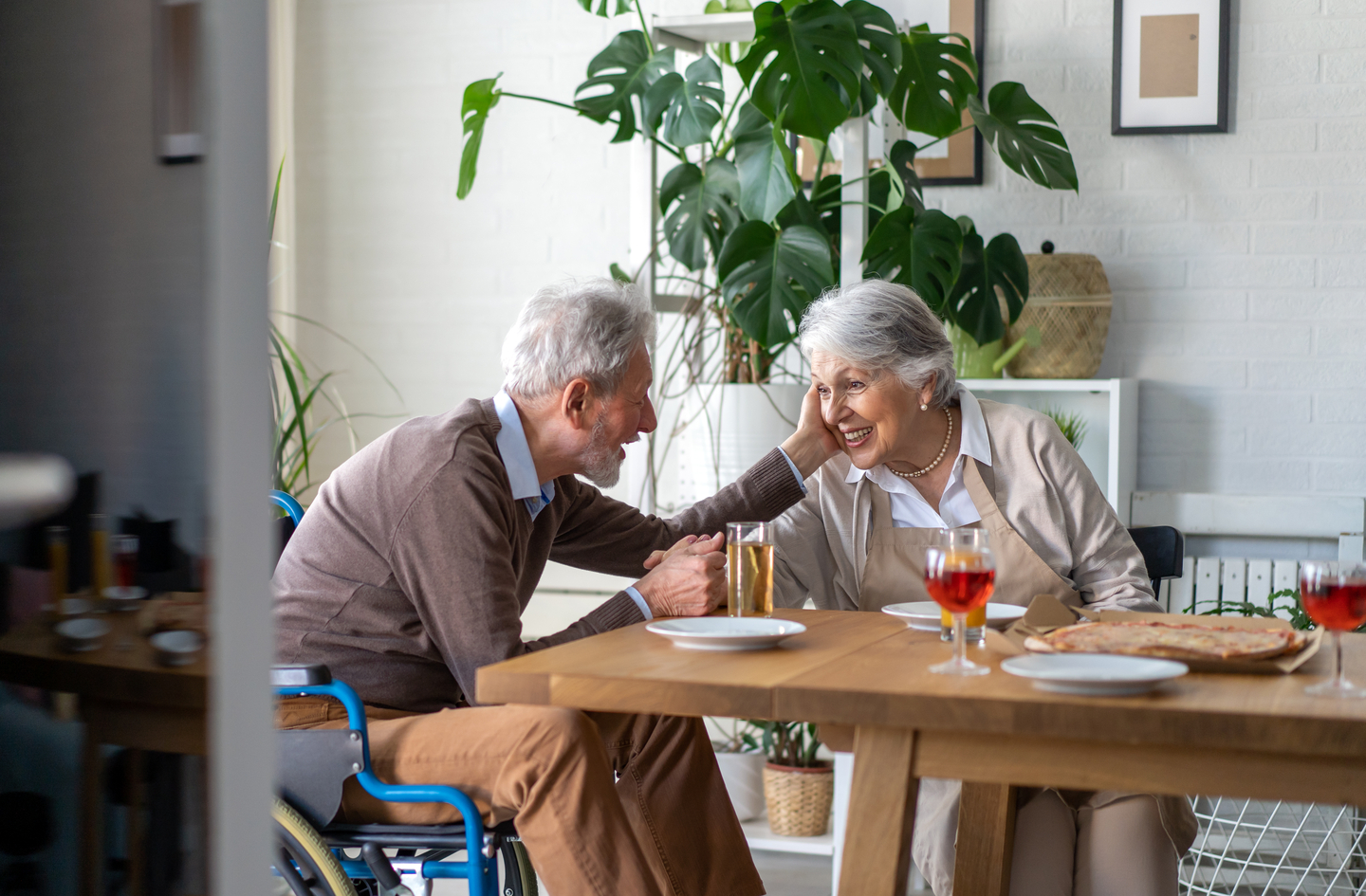An older man in a wheelchair and his wife share a meal together at a dining table in a bright, plant-filled home kitchen setting