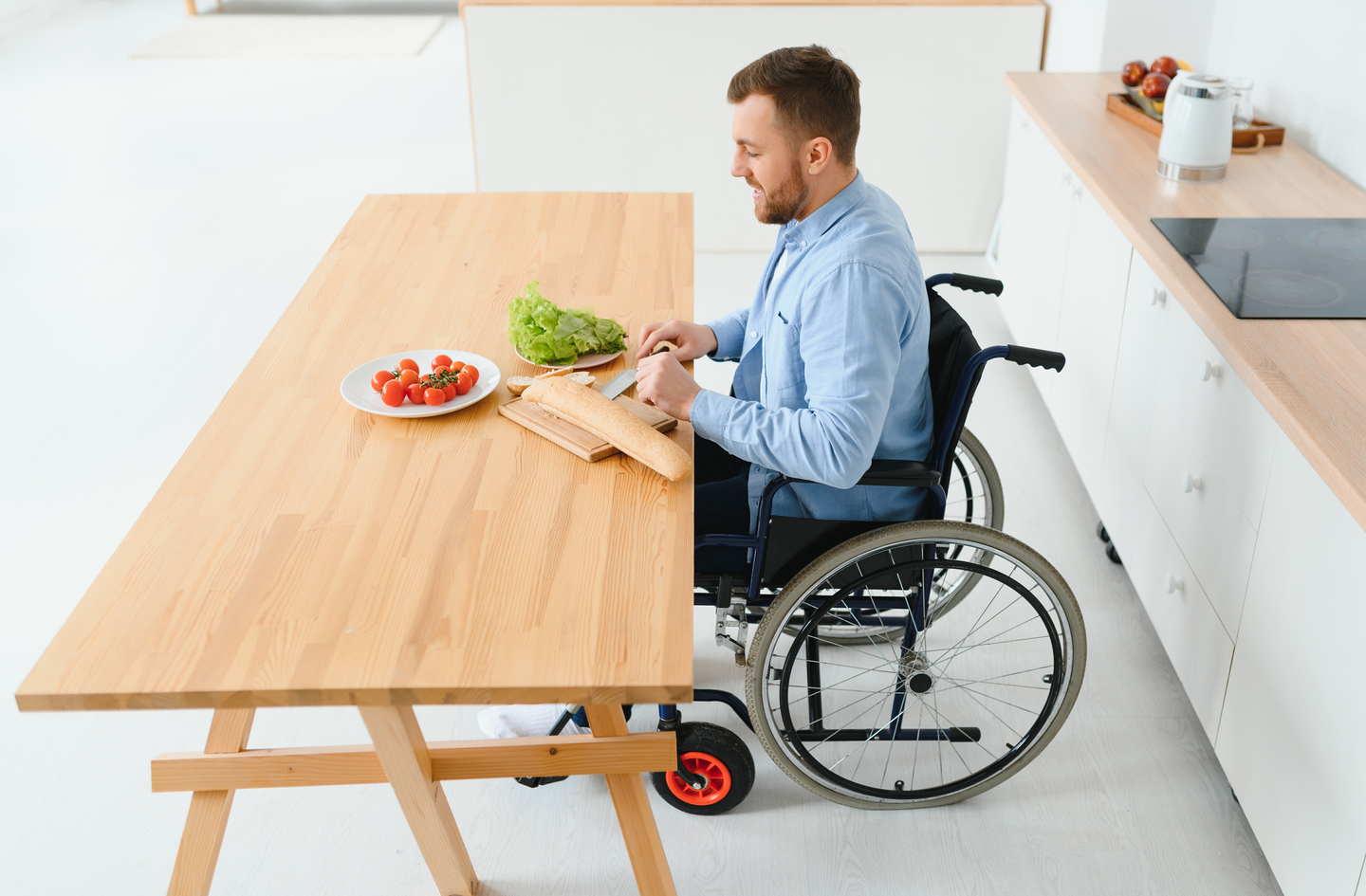 A man in a wheelchair prepares a meal at a wooden kitchen table, demonstrating reach and body position at a lowered work surface