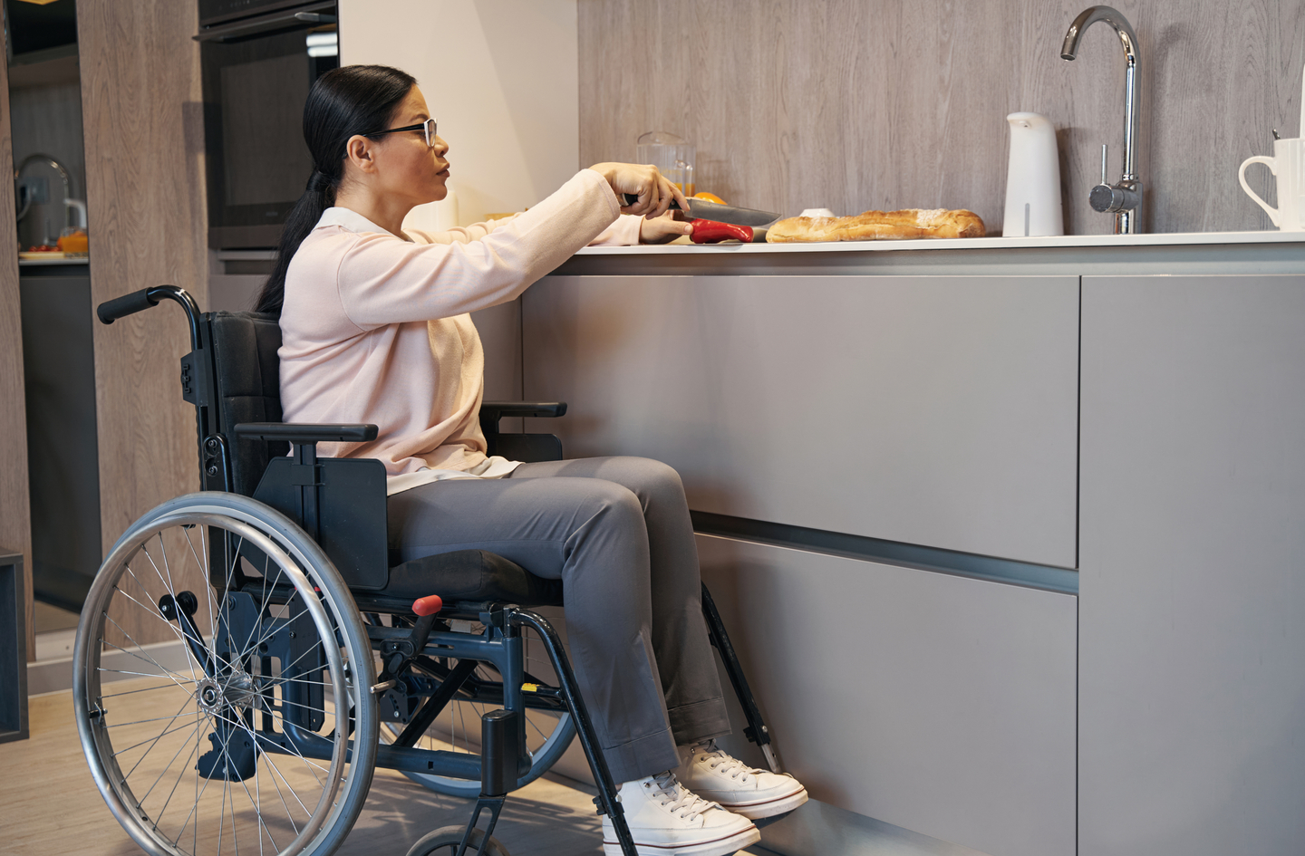 wheelchair accessible counter height demonstrated as a woman in a wheelchair cuts a red pepper at a kitchen counter, reaching forward comfortably