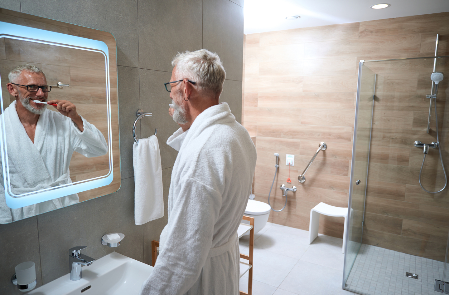 An older man brushes his teeth in a modern bathroom featuring a walk-in shower with safety grab bars and a shower stool