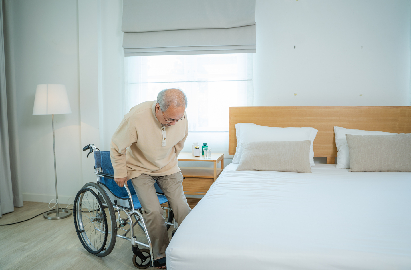 An older adult using a bed rail for the elderly to support a wheelchair-to-bed transfer beside a wooden nightstand