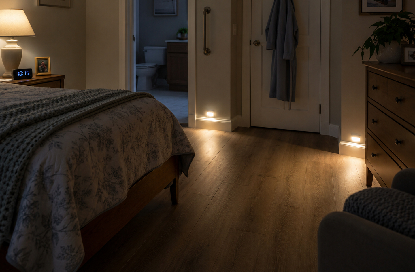 A bedroom at night with a glowing lamp, nightlights near the floorboards, and an open doorway to a bathroom - the environment surrounding a bedside commode for elderly nighttime use