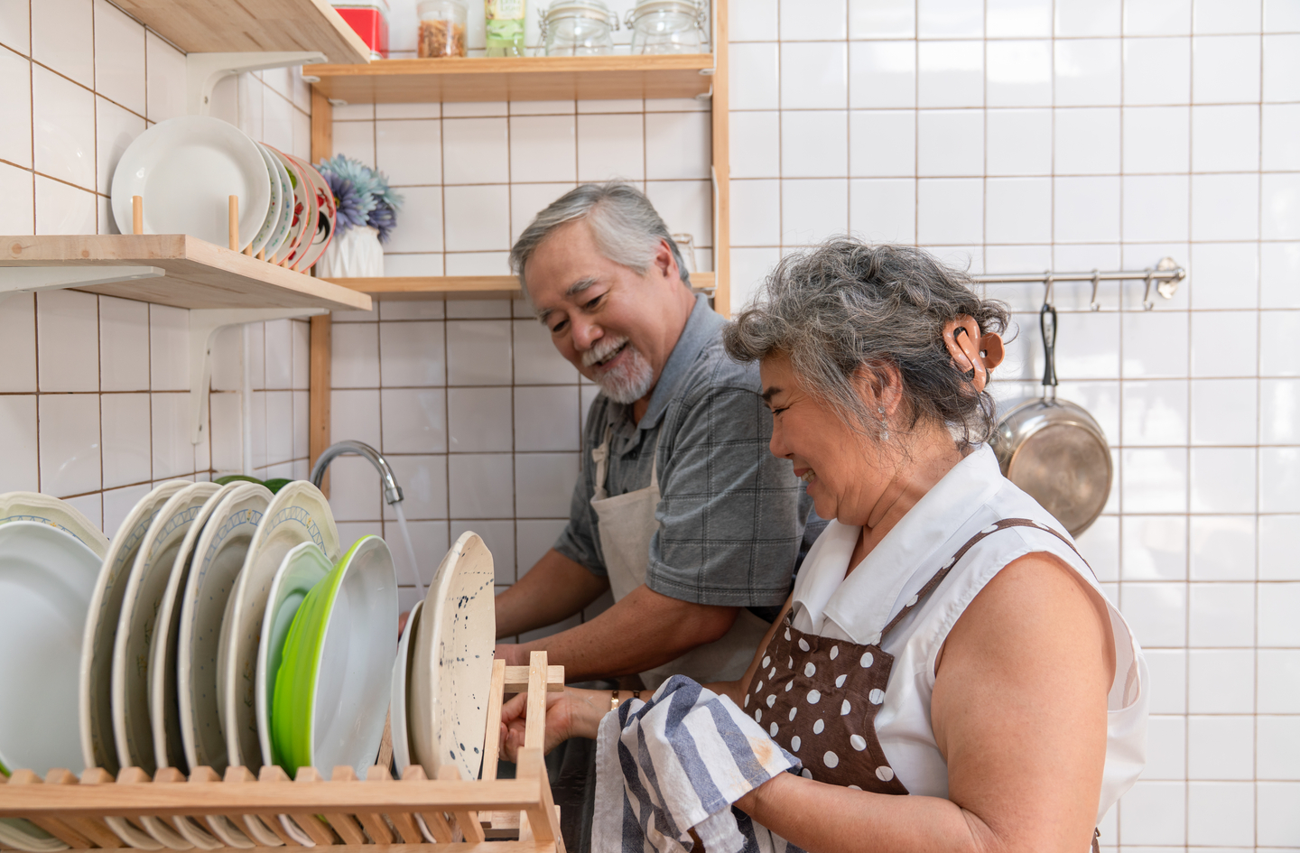 An elderly couple smiling while working together in a kitchen, with the man washing dishes at the sink and the woman drying them next to white-tiled walls with wooden shelves