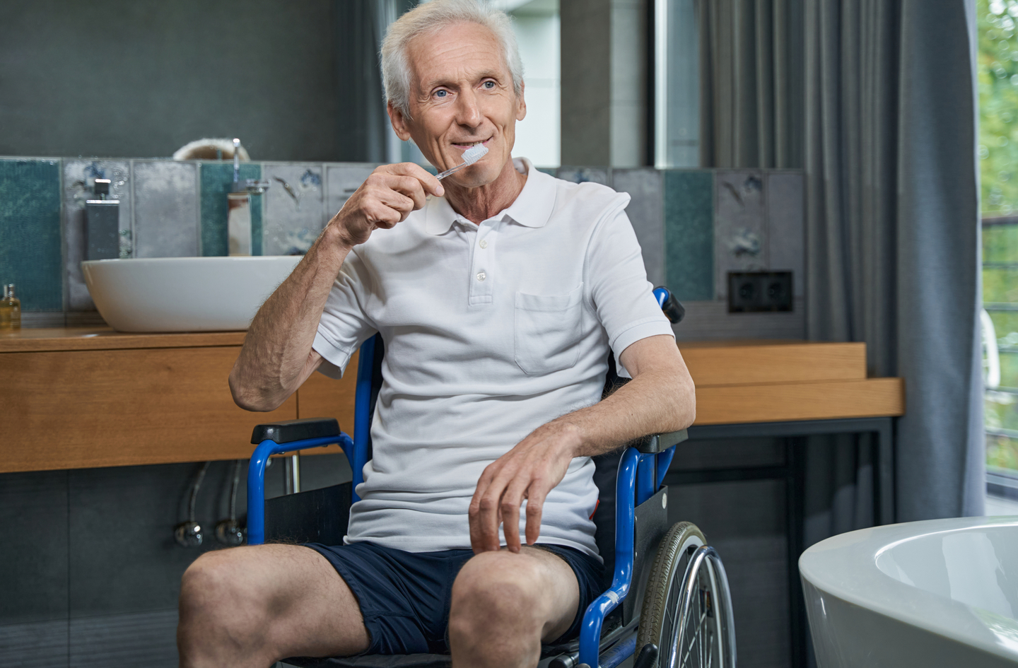 An older man in a wheelchair brushes his teeth at a bathroom sink, illustrating accessible bathroom ideas for seniors who use mobility aids