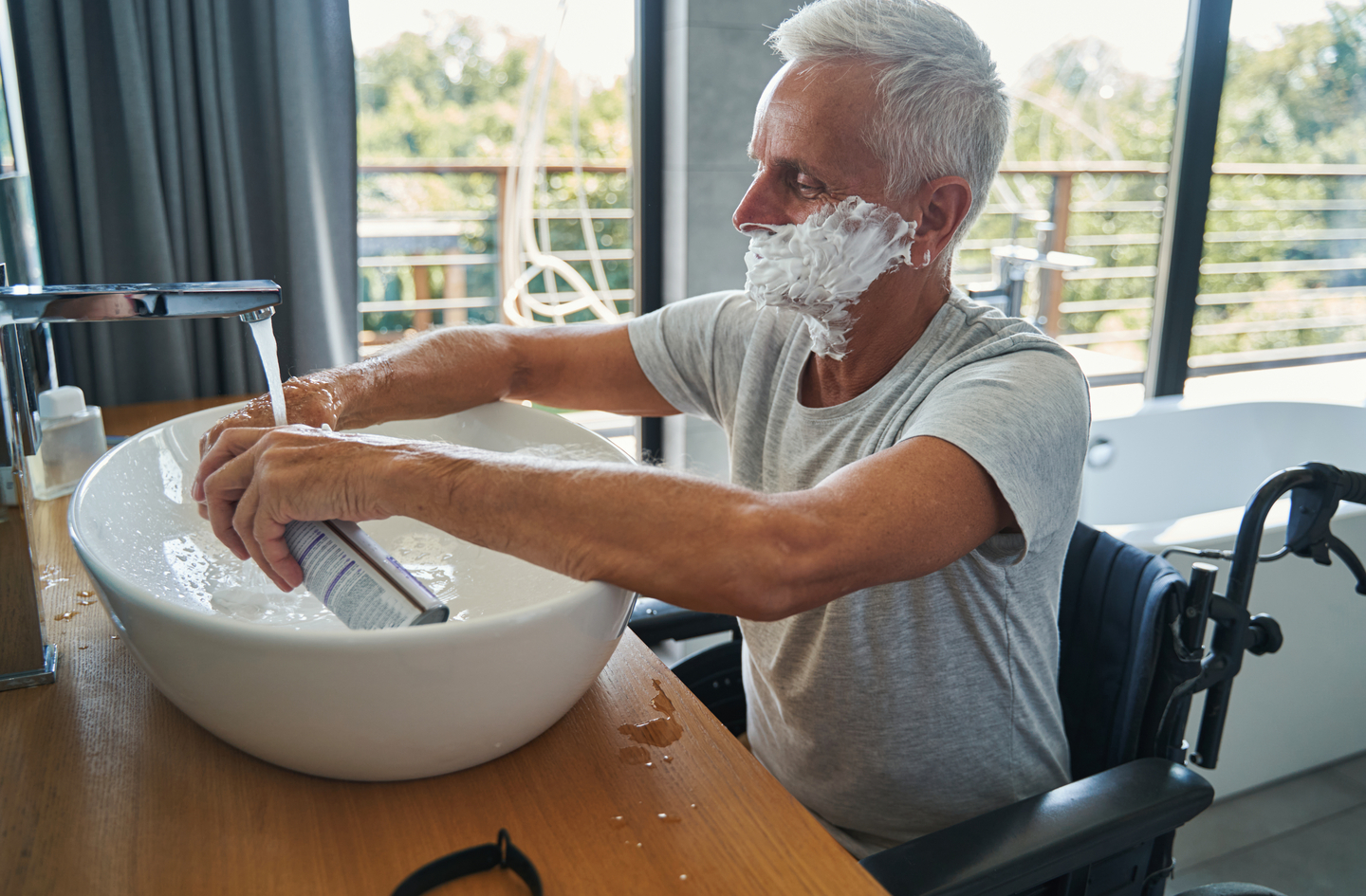 A man in a wheelchair using handicap bathroom fixtures at a sink in a modern, well-lit accessible bathroom