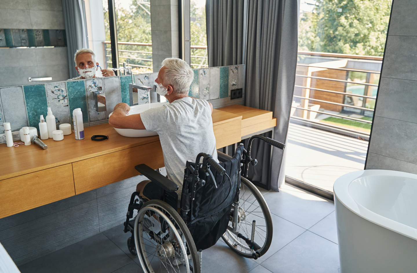 A man in a wheelchair at a bathroom vanity in a tiled bathroom, demonstrating accessible sink approach space within handicap bathroom dimensions