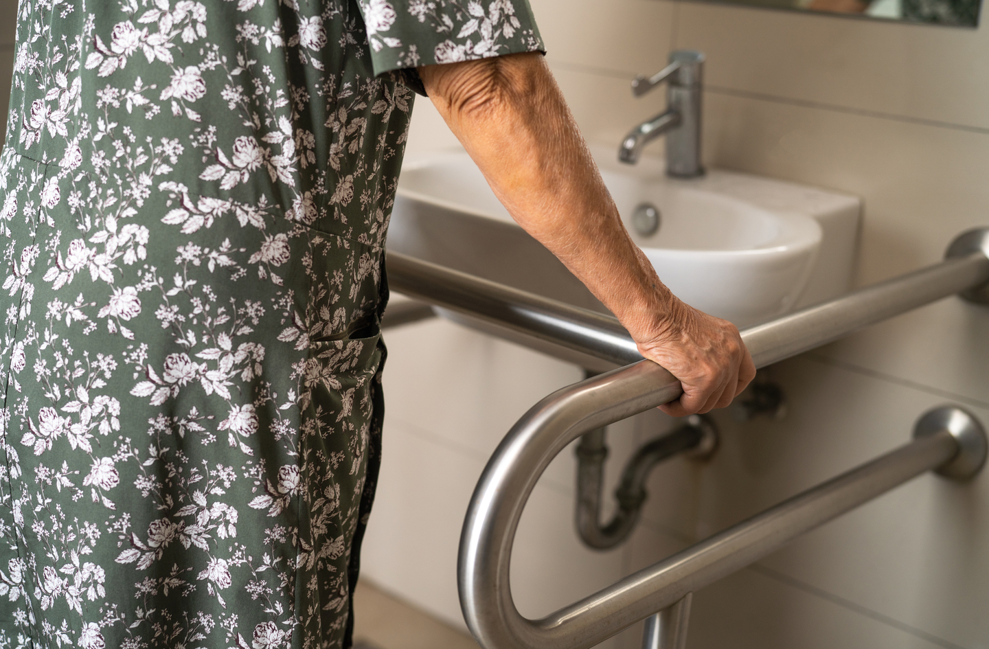 An elderly person wearing a floral green dress leans on a metal safety rail next to a bathroom sink. The person's hand is visible grasping the sturdy support bar while they stand in the tiled room.