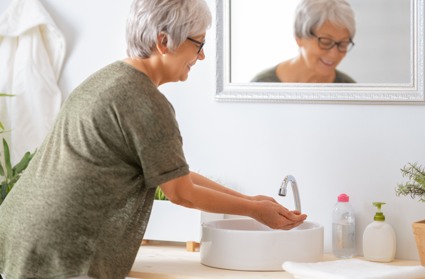 A woman with short gray hair and glasses smiles while washing her hands at a bright bathroom sink - the bathroom appears on every home safety checklist for seniors as one of the three highest-risk rooms