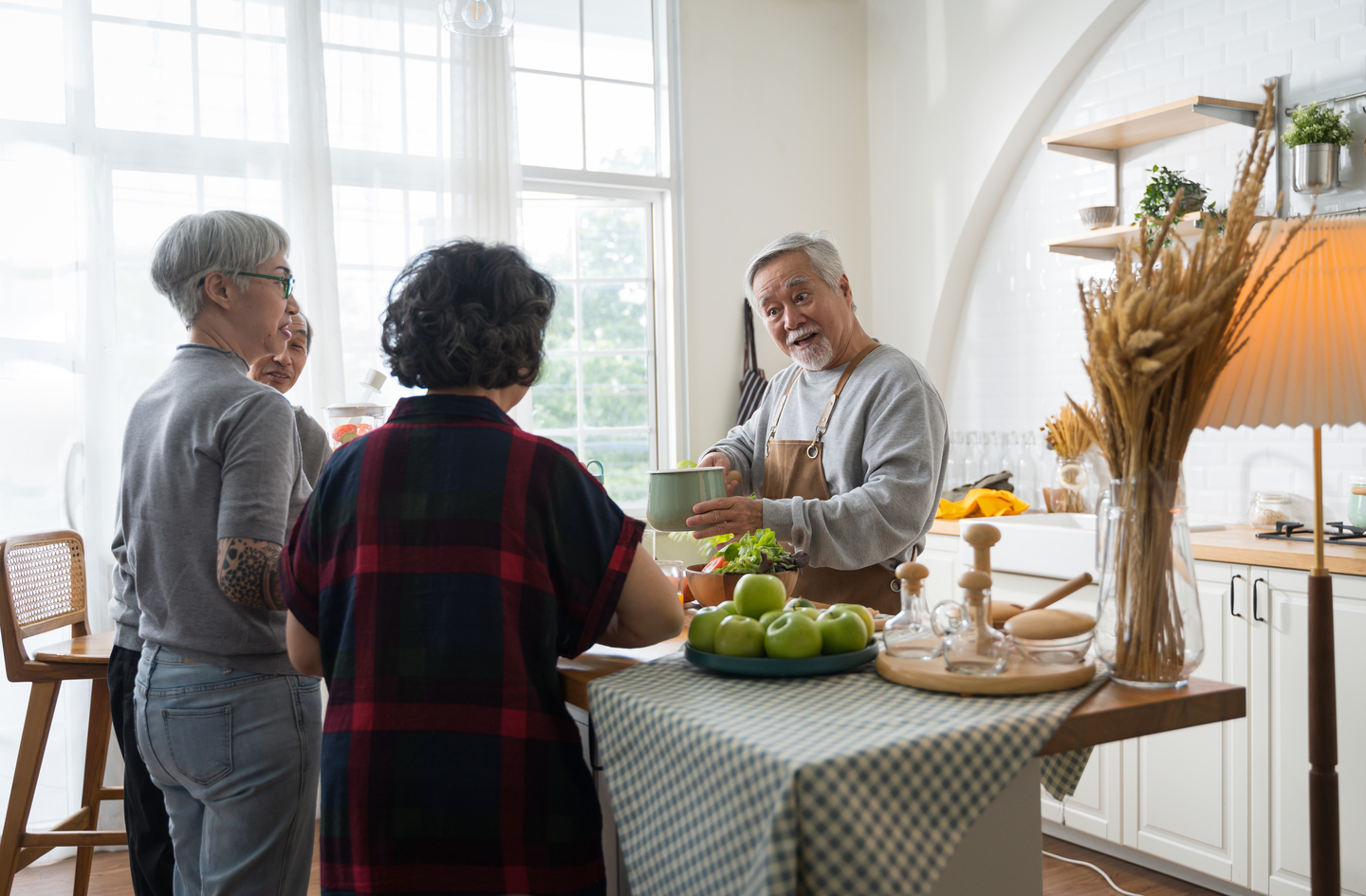 An older man wearing an apron prepares food alongside two women in a bright, modern kitchen with open counters and overhead lighting