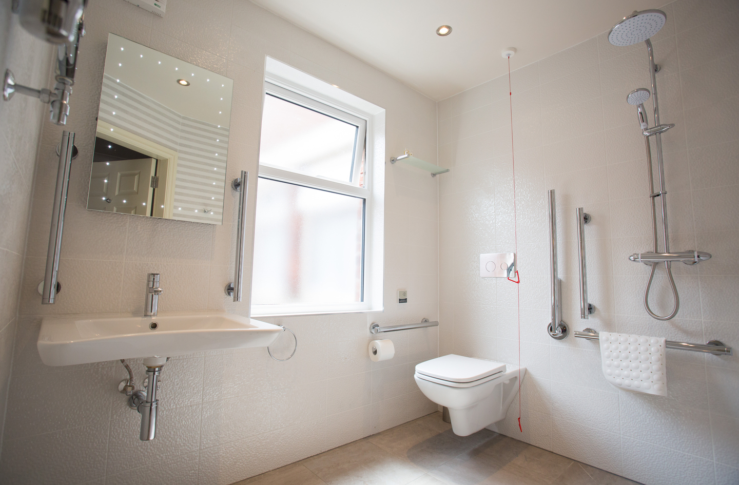 A modern accessible bathroom with white tiled walls, a wall-mounted toilet with safety grab bars, a vanity sink, and an emergency alarm pull cord - the kind of support features a home safety checklist for seniors identifies in this room