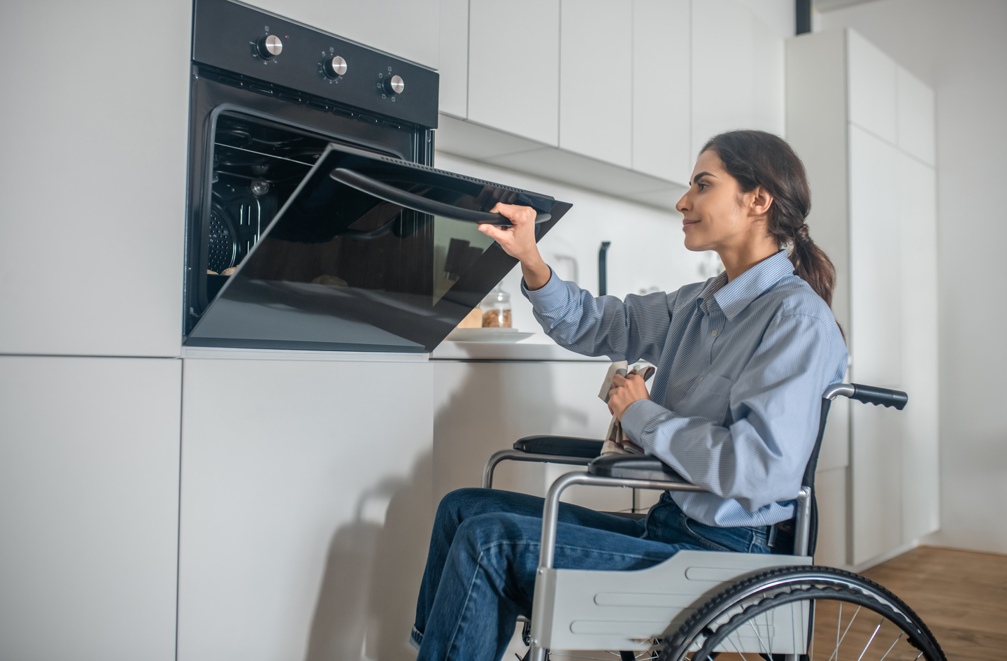 A woman in a wheelchair opens a built-in oven door in a modern white kitchen, showing accessible appliance placement at a reachable height.