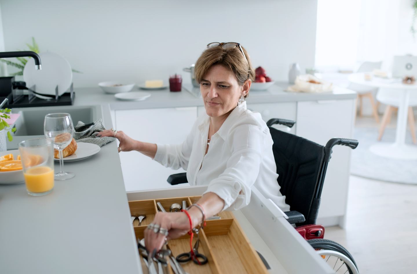 A woman in a wheelchair reaching into an open kitchen drawer to retrieve silverware, demonstrating accessible drawer storage at a comfortable height
