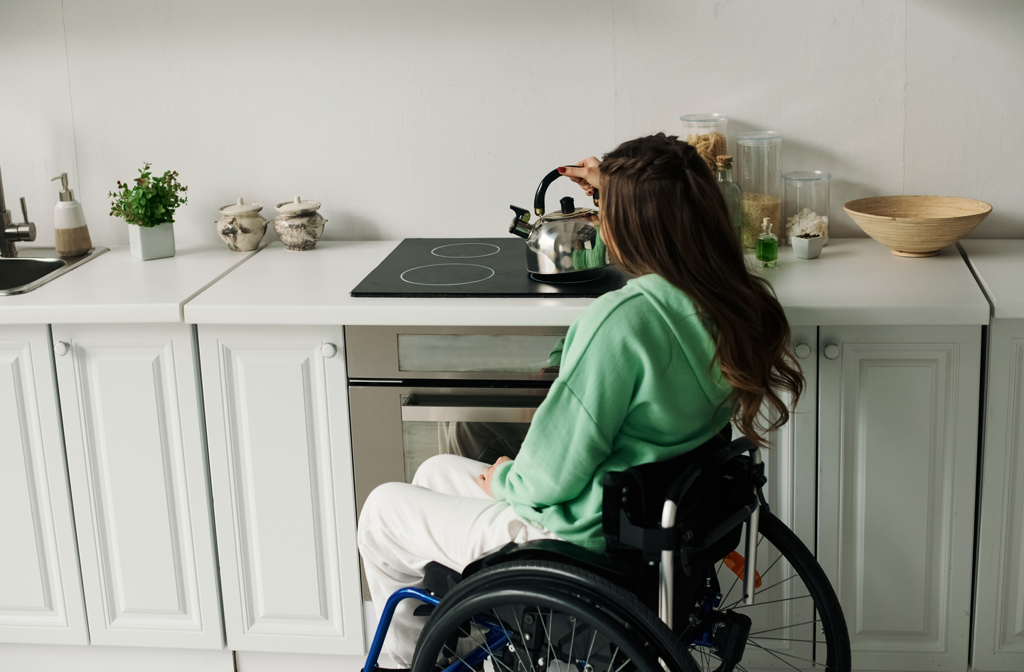 A person in a wheelchair reaching toward a kitchen stovetop, demonstrating why reach range and counter height are central to accessible kitchen requirements
