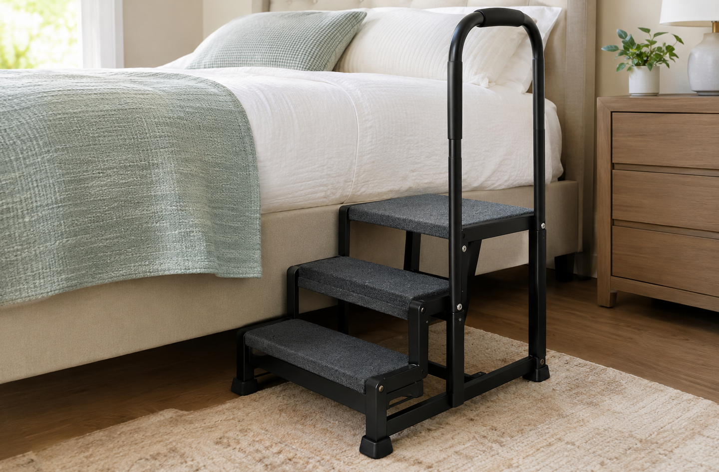 A three-step bedside staircase with an integrated handrail positioned beside a bed, showing gray-textured step treads and a black frame resting on a bedroom rug.