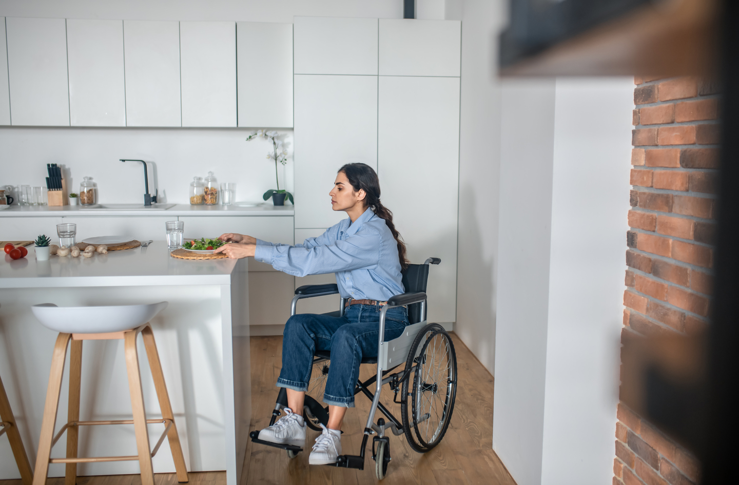 A young woman in a wheelchair sits at a white kitchen island preparing a salad, with ingredients within easy reach - a working example of accessible kitchen design.