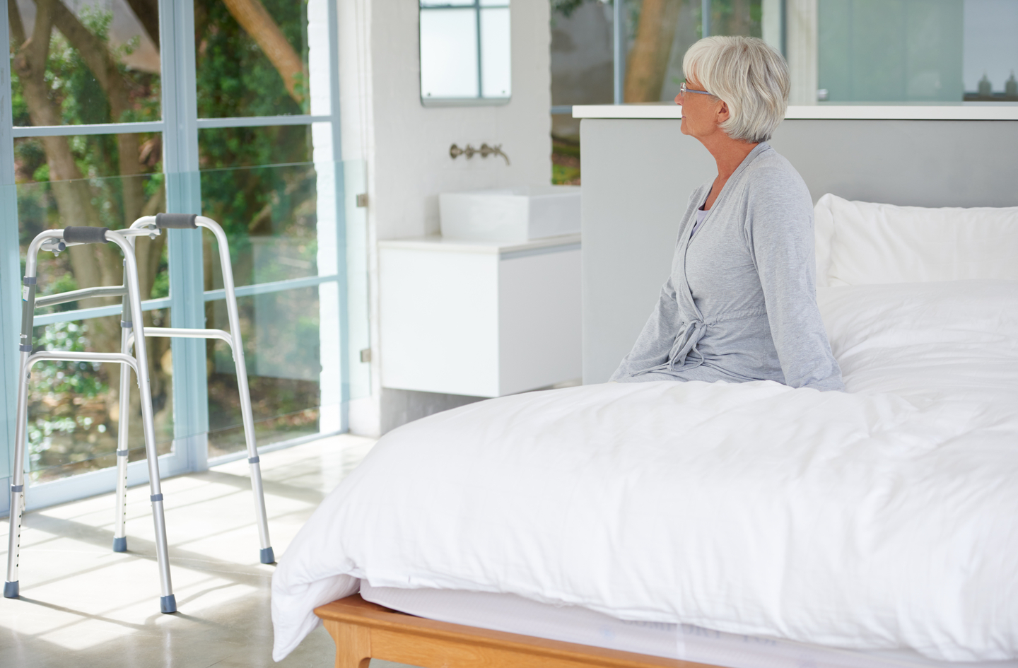 An older adult sits on the edge of a bed in a modern bedroom while a metal walker stands on the floor nearby, showing the typical positioning of a mobility aid in the bed-to-floor transfer zone where a rug would be placed.