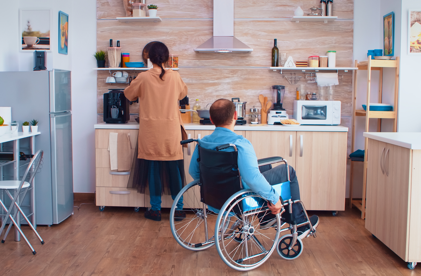 Accessible kitchen design in use: a woman stands at a counter while a man in a wheelchair works beside her in a well-lit kitchen with light wood cabinets.