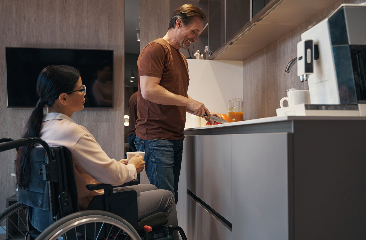 A man prepares food at a kitchen counter while a woman using a wheelchair sits beside him holding a cup, in a modern kitchen with neutral tones.