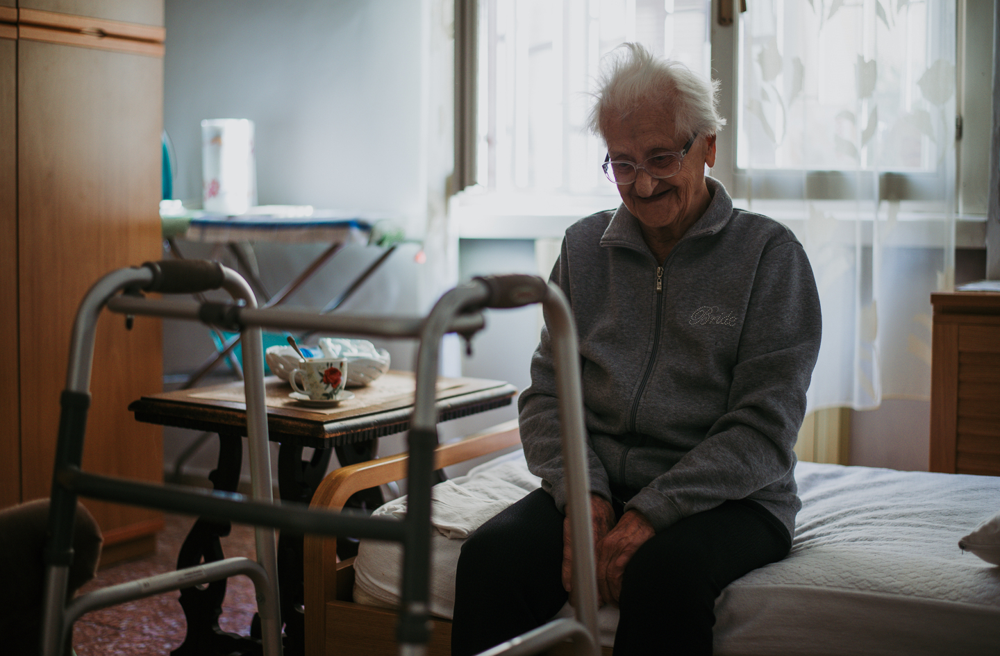 Senior bedroom safety: an older woman with white hair and glasses smiles while seated on her bed, with a walker parked within reach and a small bedside table behind it