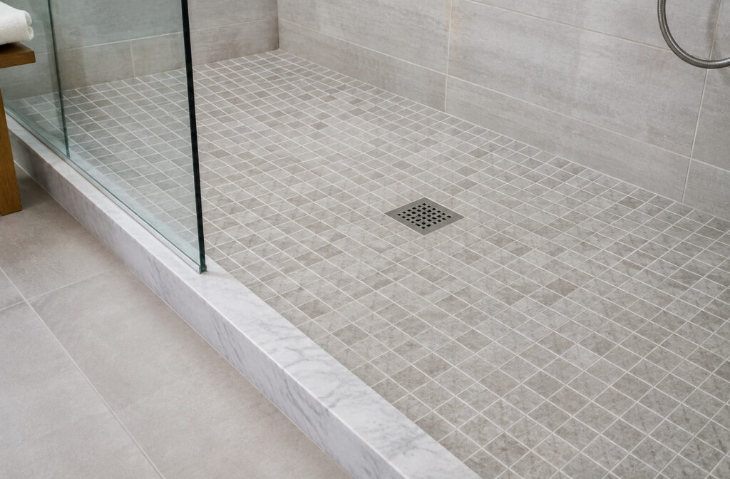 Slip-resistant flooring in a walk-in shower: a 2-inch grid of gray textured tiles around a centered square metal drain, with a glass panel and a marble threshold